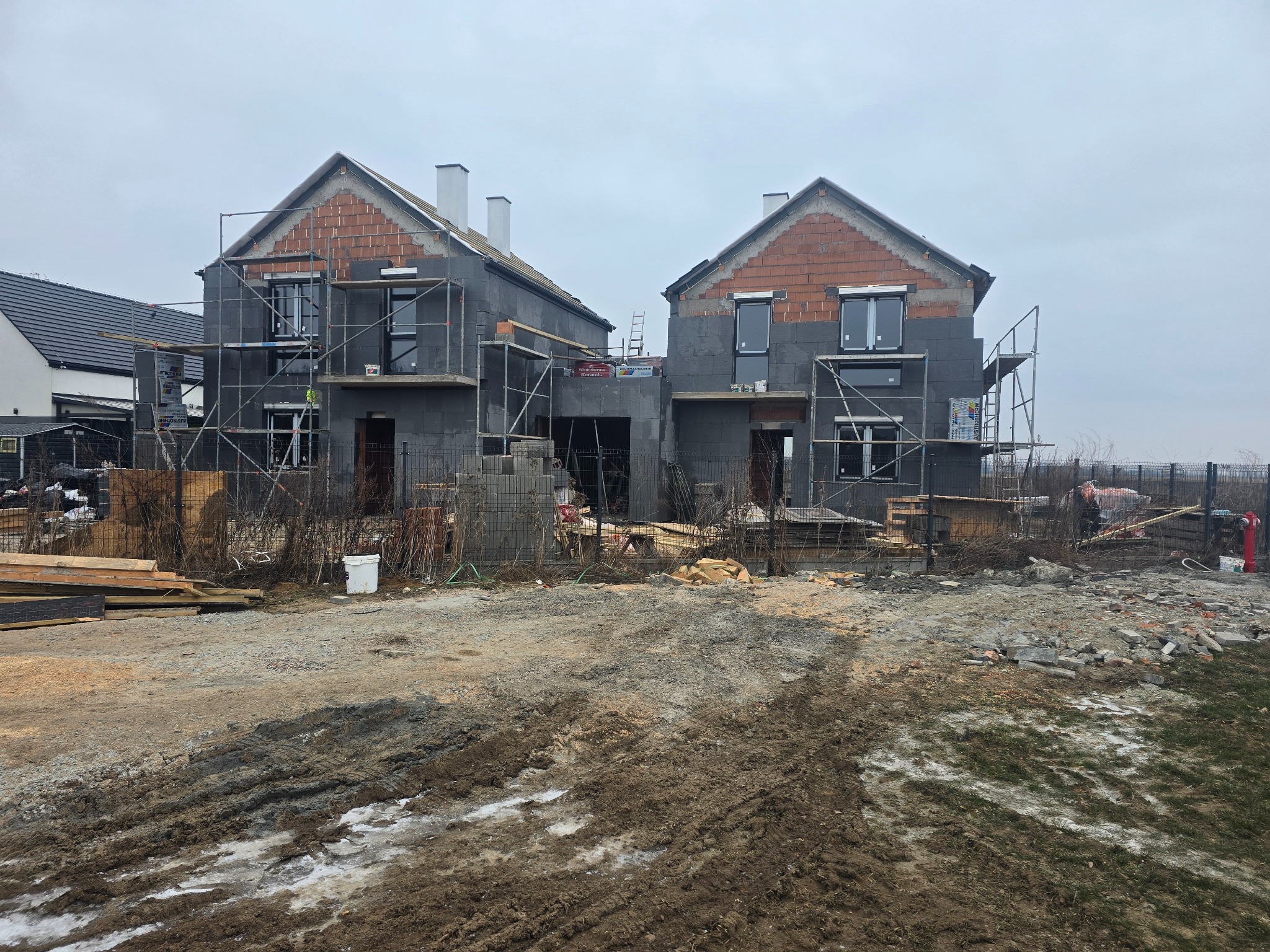 Dwa domy w stanie surowym zamkniętym, z szarymi ścianami i ceglanymi szczytami, otoczone rusztowaniami i materiałami budowlanymi na błotnistym terenie.