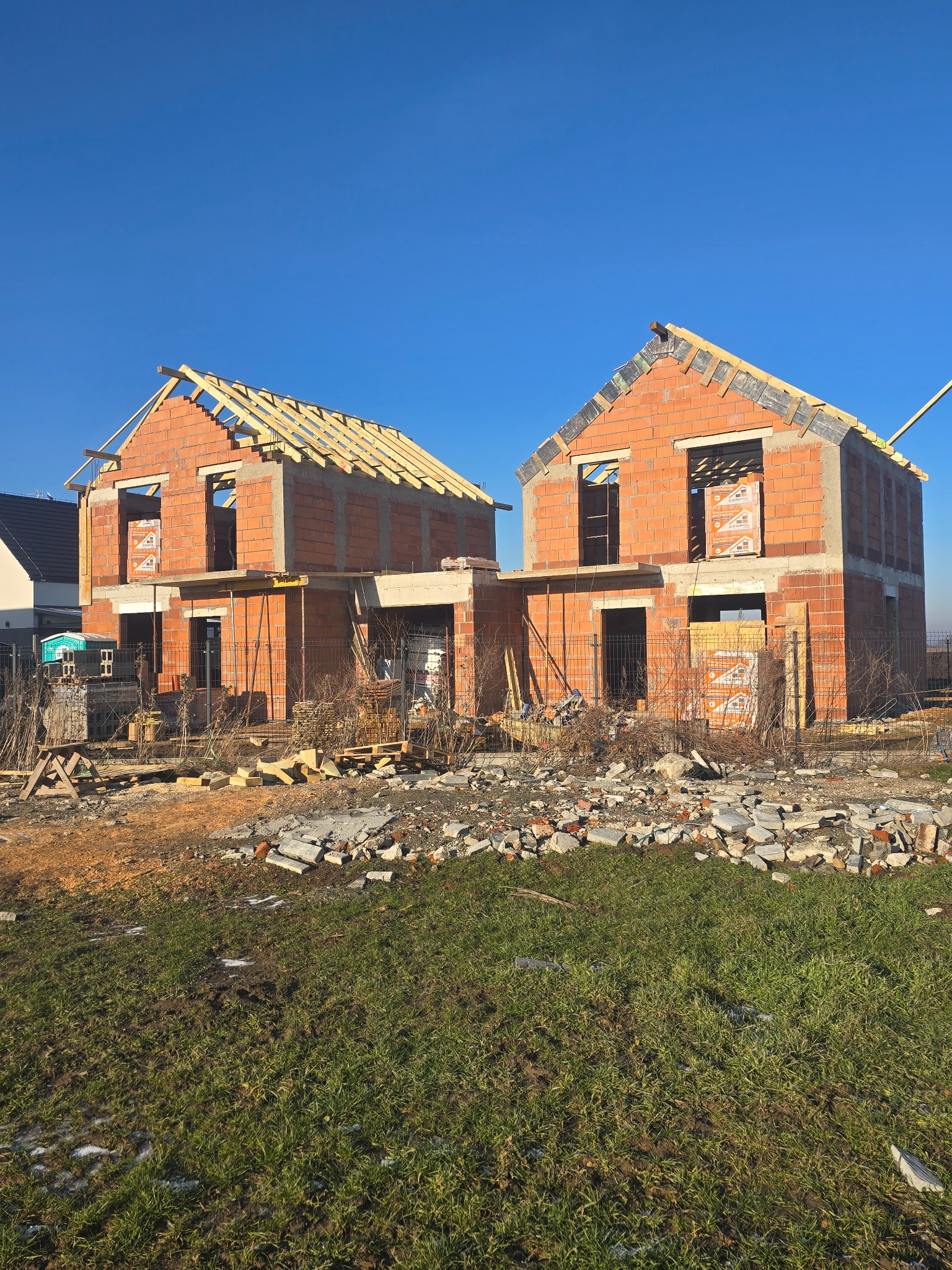 Dwa domy w stanie surowym otwartym, z czerwonej cegły, z drewnianą konstrukcją dachu, na tle błękitnego nieba. Widoczne rusztowania i materiały budowlane.