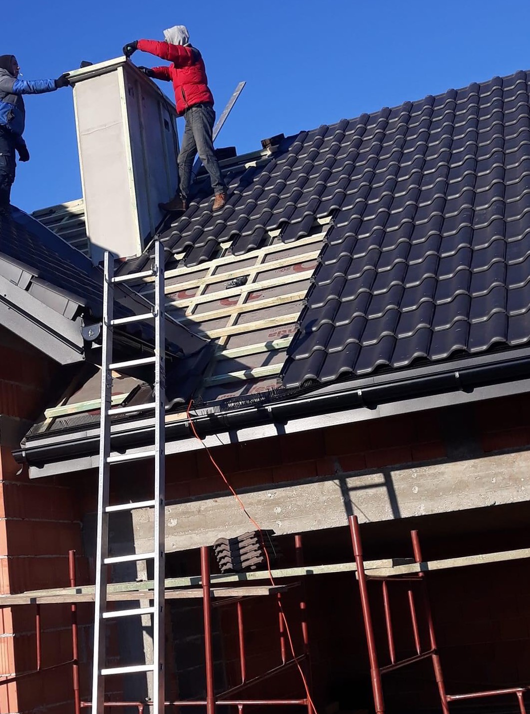 Dwóch pracowników na dachu w trakcie montażu komina wentylacyjnego, widoczna konstrukcja dachu z łatami i częściowo ułożona dachówka ceramiczna w kolorze grafitowym.