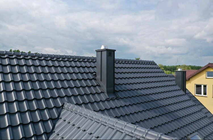 Nowy, ciemnoszary dach z blachodachówki z dwoma kominami na tle pochmurnego nieba i fragmentu sąsiedniego domu.