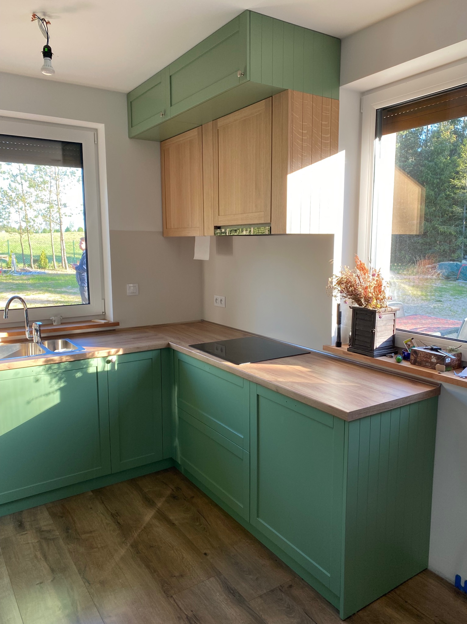 Kąt ujęcia na zieloną kuchnię z drewnianymi blatami i szafkami wiszącymi w odcieniach drewna i zieleni, widok na zewnątrz przez okno, dekoracje na blacie.