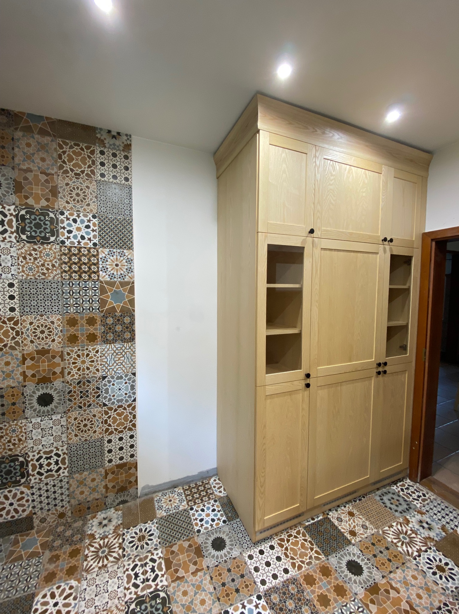 Wysoka, jasna szafa z drewna z przeszklonymi górnymi szafkami, dopasowana do wzorzystych płytek na ścianie i podłodze w stylu marokańskim.