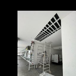 MATEUSZ MOTOR - Pracownik w kombinezonie na drabinie przy ażurowym otworze w suficie podczas prac wykończeniowych w lokalu usługowym, widoczne rusztowanie i samochód dostawczy.