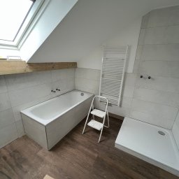 MATEUSZ MOTOR - Łazienka w trakcie remontu: biała wanna obudowana płytkami, kabina prysznicowa, grzejnik drabinkowy, drewniana belka pod oknem dachowym, brązowe panele podłogowe, drabina malarska i kartony...