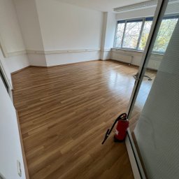 MATEUSZ MOTOR - Puste biuro z drewnianą podłogą, białymi ścianami i oknami z widokiem na drzewa, z czerwonym gaśnicą przy drzwiach.