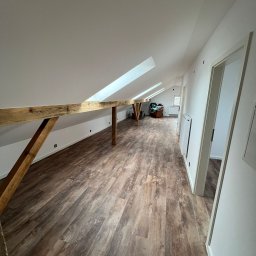 MATEUSZ MOTOR - Poddasze po remoncie z drewnianą podłogą imitującą panele, białymi ścianami, drewnianą belką konstrukcyjną i oknami dachowymi.