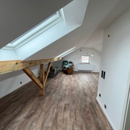 MATEUSZ MOTOR - Poddasze po remoncie z drewnianą konstrukcją nośną, oknami dachowymi i panelami podłogowymi imitującymi drewno. Widoczne gniazdka elektryczne w ścianie i częściowo wyposażone wnętrze.
