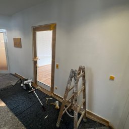 MATEUSZ MOTOR - Korytarz w trakcie malowania: ściany zabezpieczone taśmą, drabina malarska, wałek i kuweta na podłodze, widoczne ślady farby na drabinie.