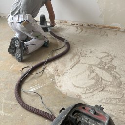 MATEUSZ MOTOR - Pracownik w Skawie szlifuje ścianę za pomocą szlifierki z odkurzaczem, przygotowując ją do dalszych prac wykończeniowych. Na podłodze widoczne ślady po szlifowaniu i odłamki starej powłoki.