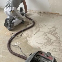 Pracownik w Skawie szlifuje ścianę za pomocą szlifierki z odkurzaczem, przygotowując ją do dalszych prac wykończeniowych. Na podłodze widoczne ślady po szlifowaniu i odłamki starej powłoki.