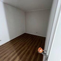 MATEUSZ MOTOR - Pusty pokój po remoncie z brązową podłogą imitującą drewno, świeżo pomalowane ściany na biało, widoczne gniazdka elektryczne i wystająca instalacja elektryczna przy futrynie drzwi.
