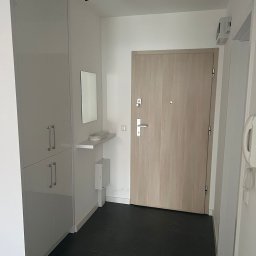 MATEUSZ MOTOR - Nowoczesny przedpokój z jasnymi szafami w zabudowie, drewnianymi drzwiami wejściowymi i czarną podłogą z płytek, oświetlony reflektorem sufitowym.