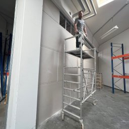 MATEUSZ MOTOR - Pracownik na rusztowaniu montuje płyty gipsowo-kartonowe na ścianie w przestrzeni magazynowej z widocznymi regałami.