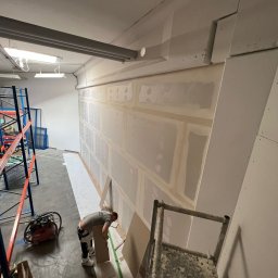 MATEUSZ MOTOR - Pracownik w szarej koszulce przykłada płytę gipsową do ściany w pomieszczeniu magazynowym, widoczny odkurzacz przemysłowy, regały i drabina malarska.