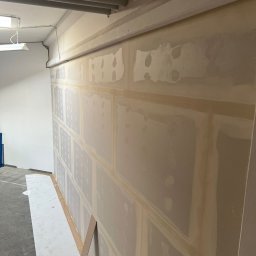 MATEUSZ MOTOR - Ściana z płyt gipsowo-kartonowych, częściowo zaszpachlowana, z widocznymi łączeniami wzmocnionymi taśmą, przygotowana do dalszych prac wykończeniowych w pomieszczeniu z betonową podłogą.