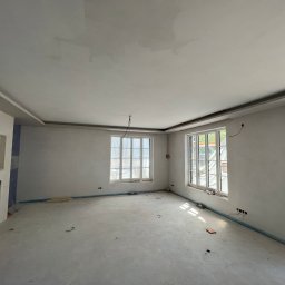 MATEUSZ MOTOR - Pokój w stanie surowym po wylewce, z przygotowanymi ścianami pod malowanie i gładzie, okna zabezpieczone folią, widoczne wystające kable elektryczne.