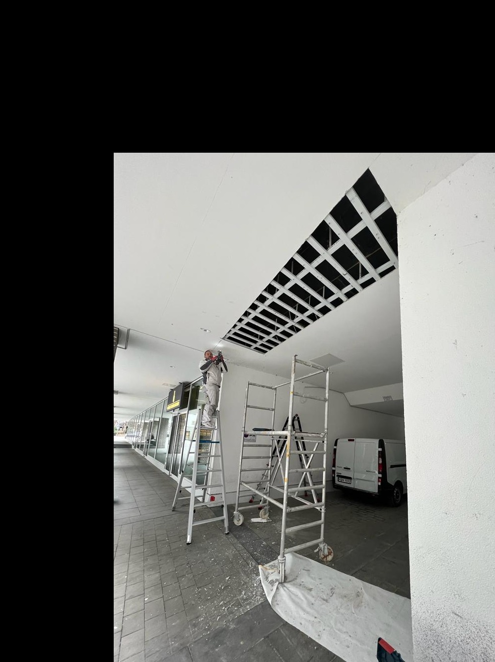 Pracownik w kombinezonie na drabinie przy ażurowym otworze w suficie podczas prac wykończeniowych w lokalu usługowym, widoczne rusztowanie i samochód dostawczy.