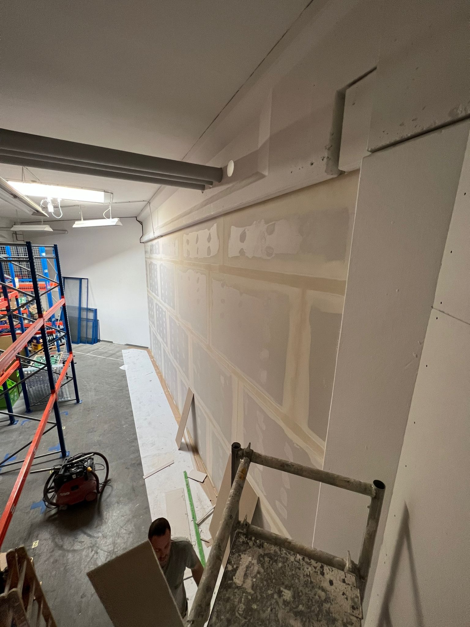 Wnętrze w trakcie prac wykończeniowych: widoczna ściana z płyt gipsowo-kartonowych z widocznymi łączeniami, drabina malarska, odkurzacz przemysłowy i fragment regału magazynowego w tle.