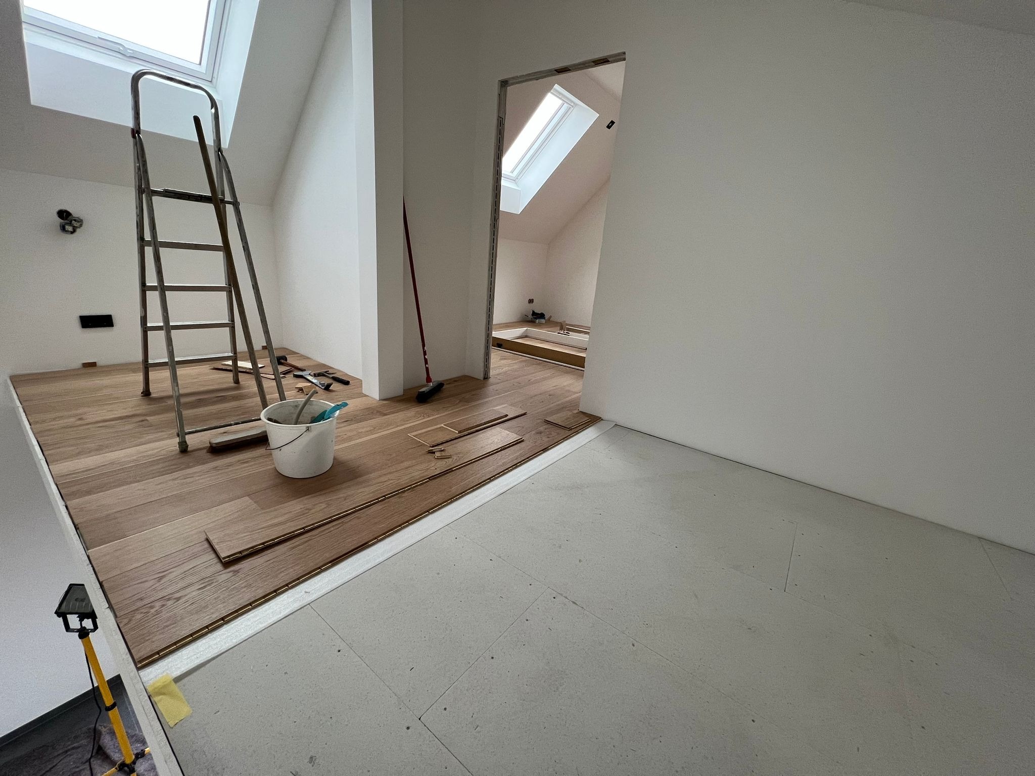 Układanie drewnianej podłogi w pokoju na poddaszu z oknem dachowym, widoczna drabina, wiadro z narzędziami i lampa budowlana.