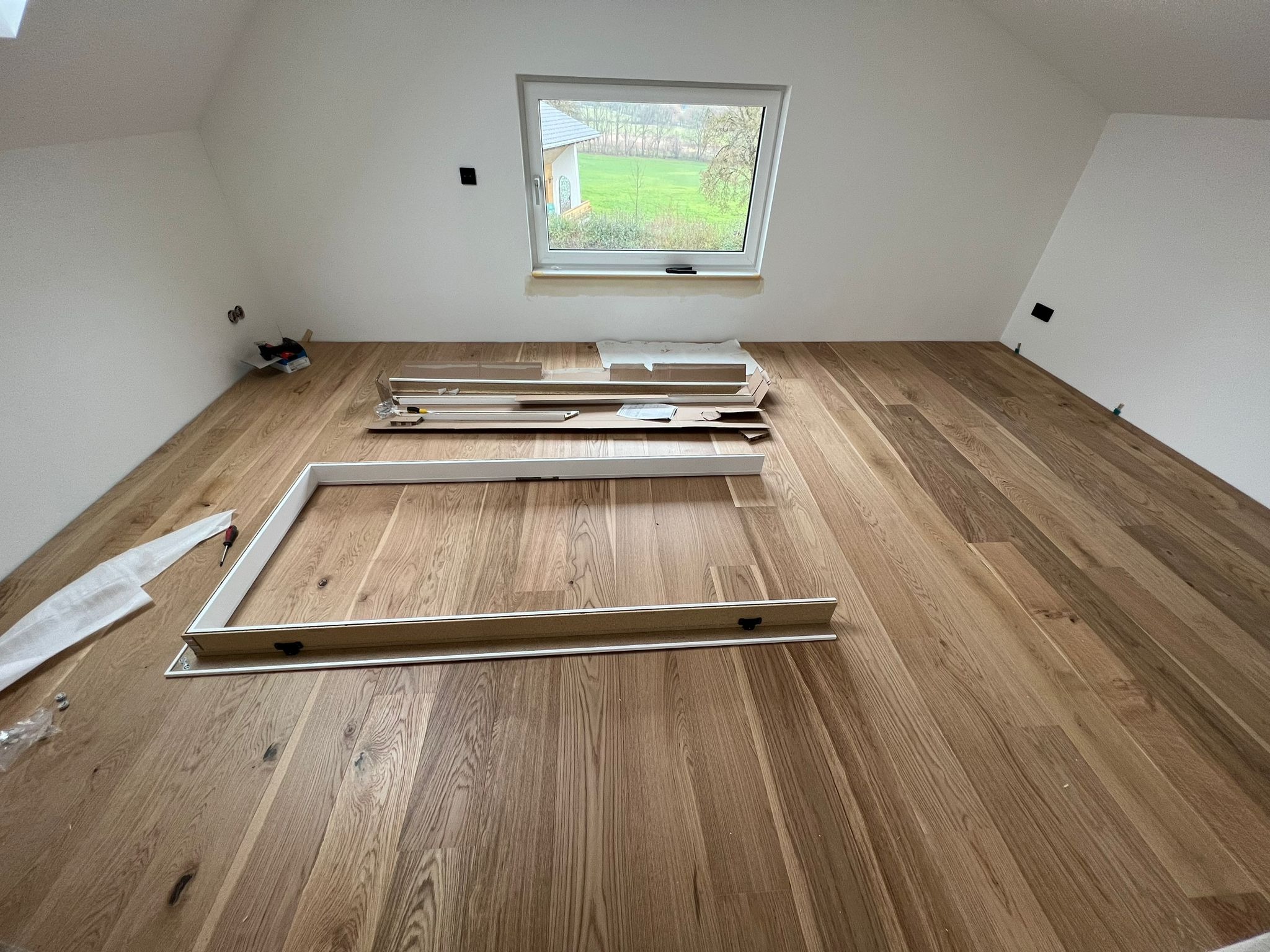 Pokój na poddaszu z drewnianą podłogą i oknem, przygotowany do montażu ościeżnicy drzwiowej. Elementy ościeżnicy leżą na podłodze obok narzędzi i opakowań.