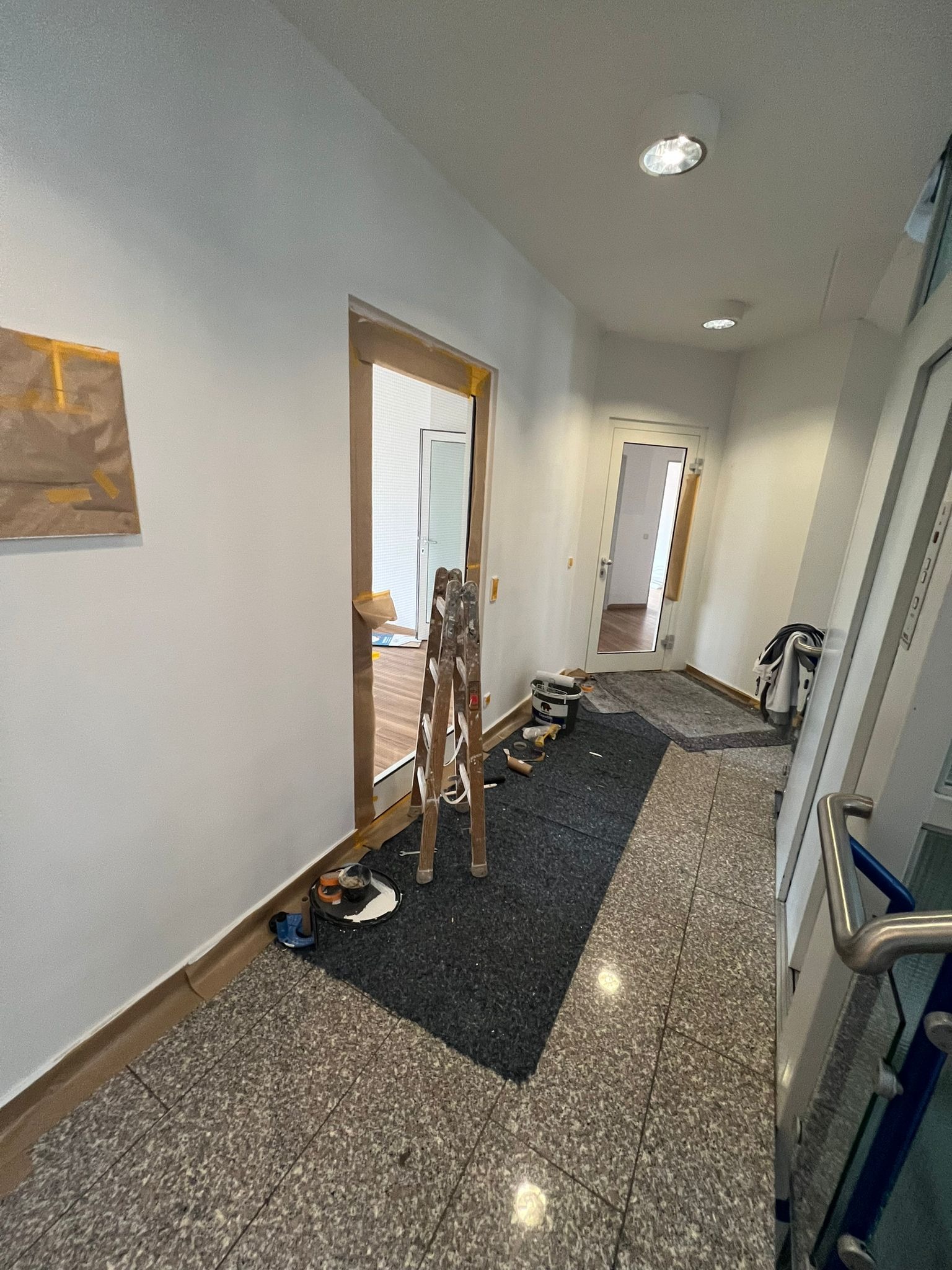 Korytarz w trakcie remontu: przygotowane ściany do malowania z widocznymi śladami taśmy malarskiej, drabina rozstawiona w przejściu, narzędzia malarskie na podłodze zabezpieczonej matą, widoczne...