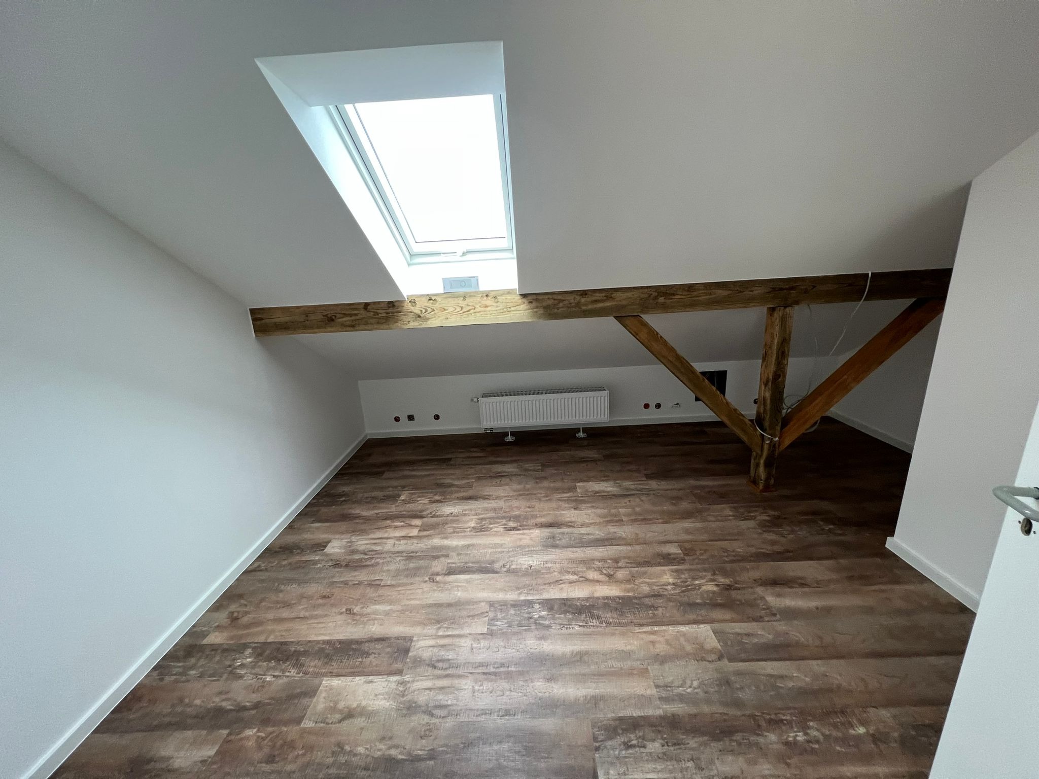 Poddasze po remoncie z drewnianą podłogą imitującą deski, białymi ścianami, drewnianą belką konstrukcyjną i oknem dachowym, widok na grzejnik i gniazdka elektryczne.
