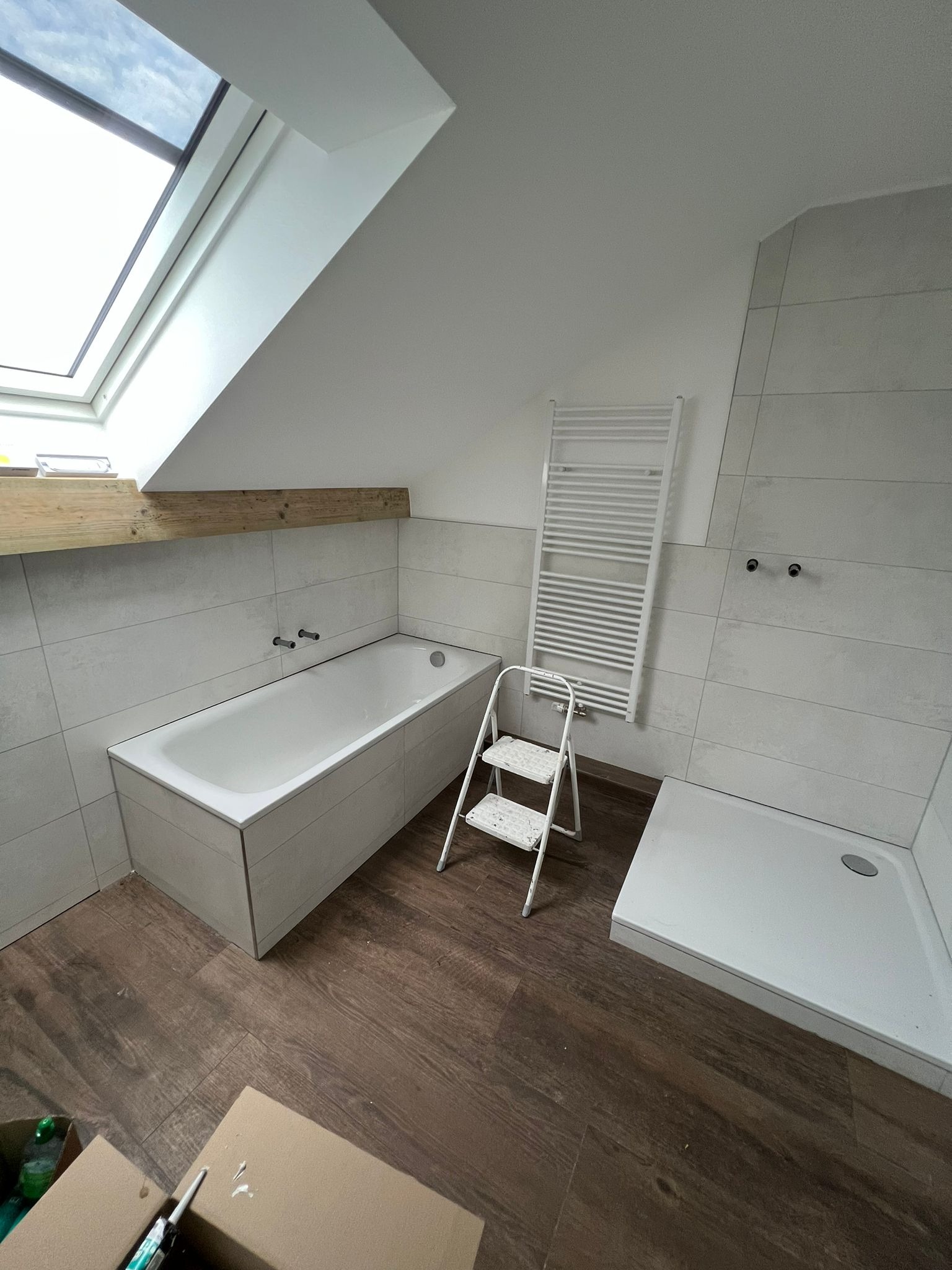 Łazienka w trakcie remontu: biała wanna obudowana płytkami, kabina prysznicowa, grzejnik drabinkowy, drewniana belka pod oknem dachowym, brązowe panele podłogowe, drabina malarska i kartony...