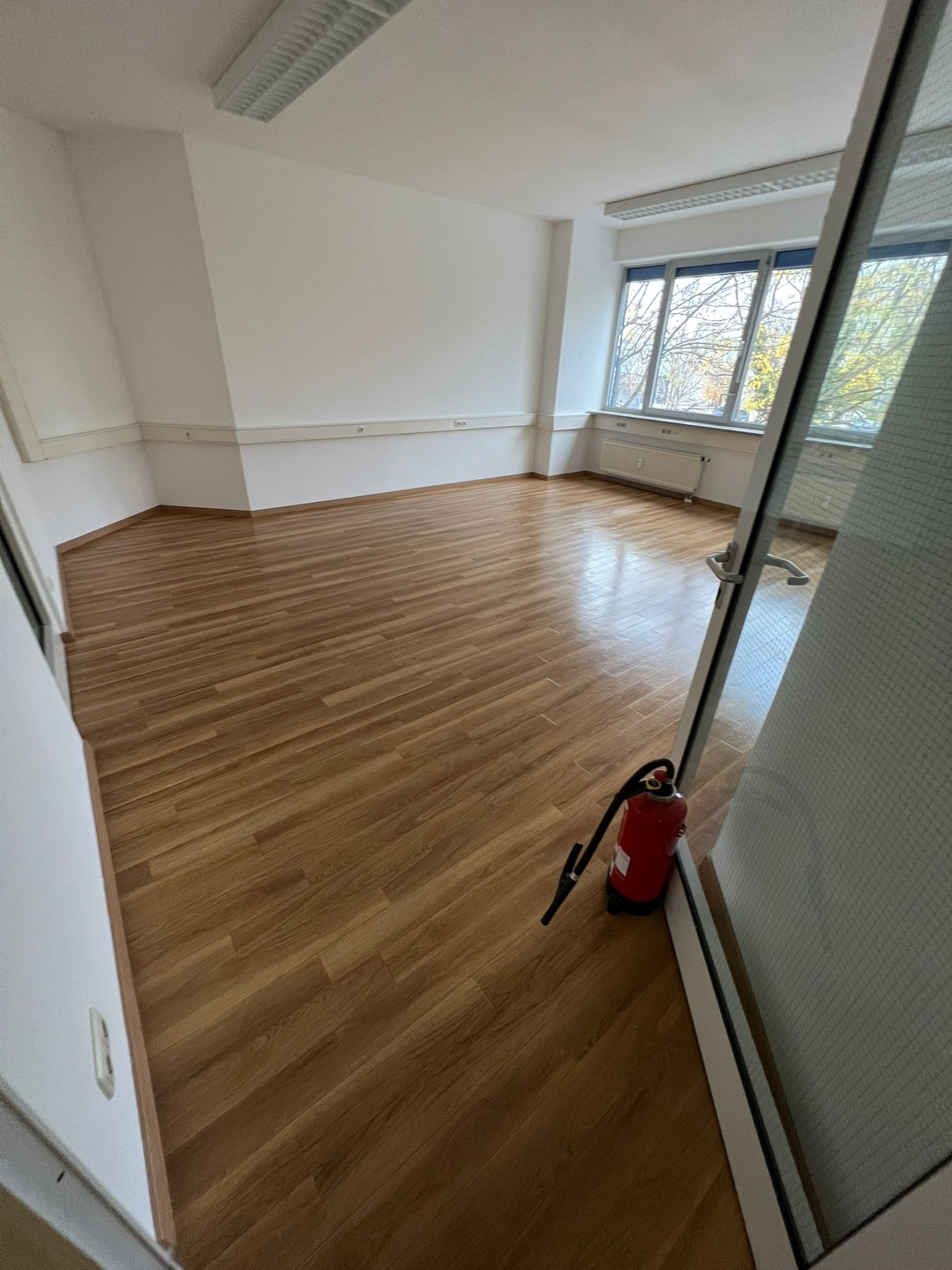 Puste biuro z drewnianą podłogą, białymi ścianami i oknami z widokiem na drzewa, z czerwonym gaśnicą przy drzwiach.