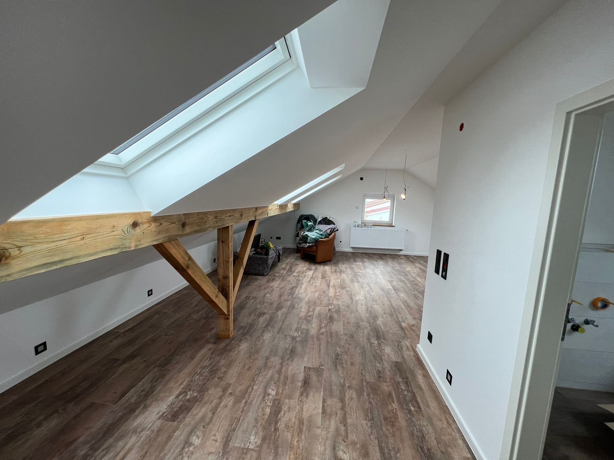 Poddasze po remoncie z drewnianą konstrukcją nośną, oknami dachowymi i panelami podłogowymi imitującymi drewno. Widoczne gniazdka elektryczne w ścianie i częściowo wyposażone wnętrze.