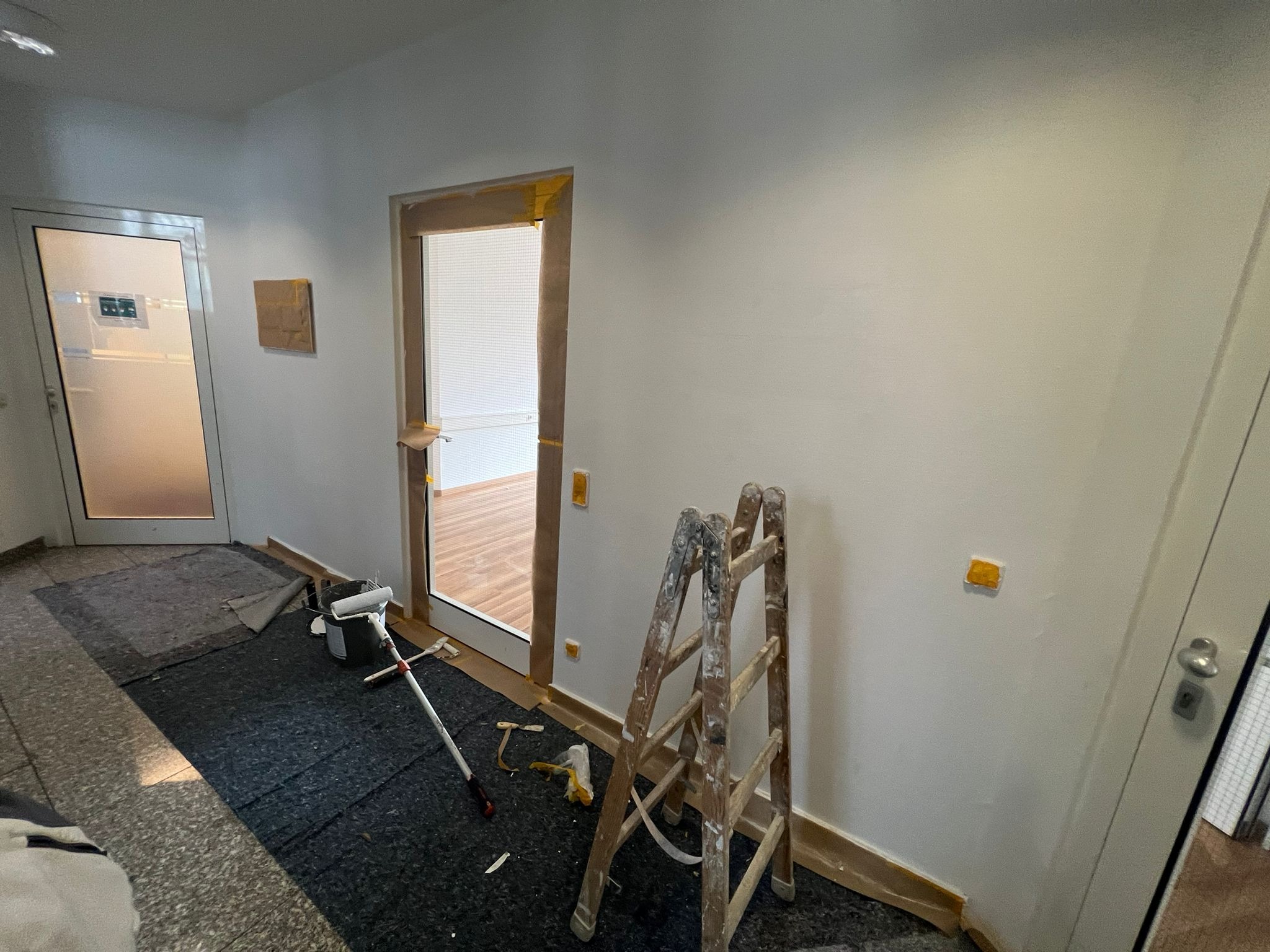 Korytarz w trakcie malowania: ściany zabezpieczone taśmą, drabina malarska, wałek i kuweta na podłodze, widoczne ślady farby na drabinie.