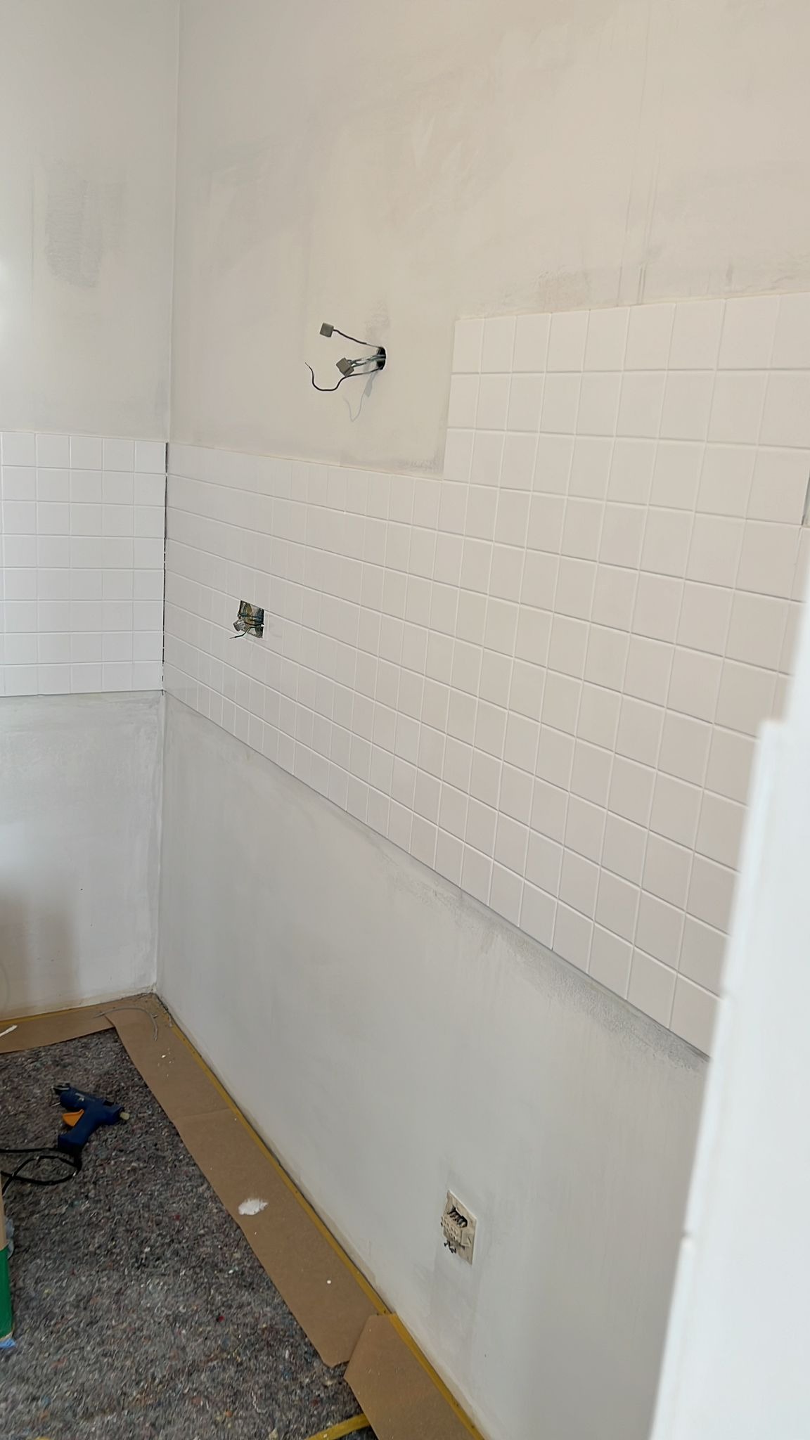 Fragment łazienki w trakcie wykańczania: białe, kwadratowe płytki częściowo ułożone na ścianie, surowa ściana powyżej, zabezpieczona podłoga i widoczne gniazdko elektryczne.