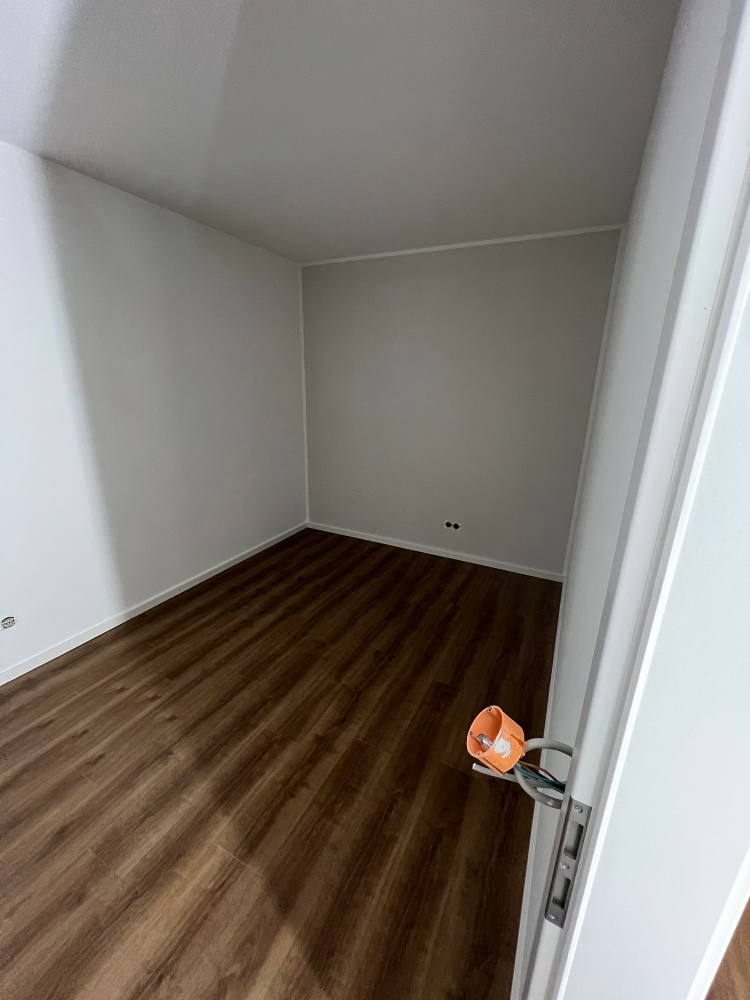 Pusty pokój po remoncie z brązową podłogą imitującą drewno, świeżo pomalowane ściany na biało, widoczne gniazdka elektryczne i wystająca instalacja elektryczna przy futrynie drzwi.