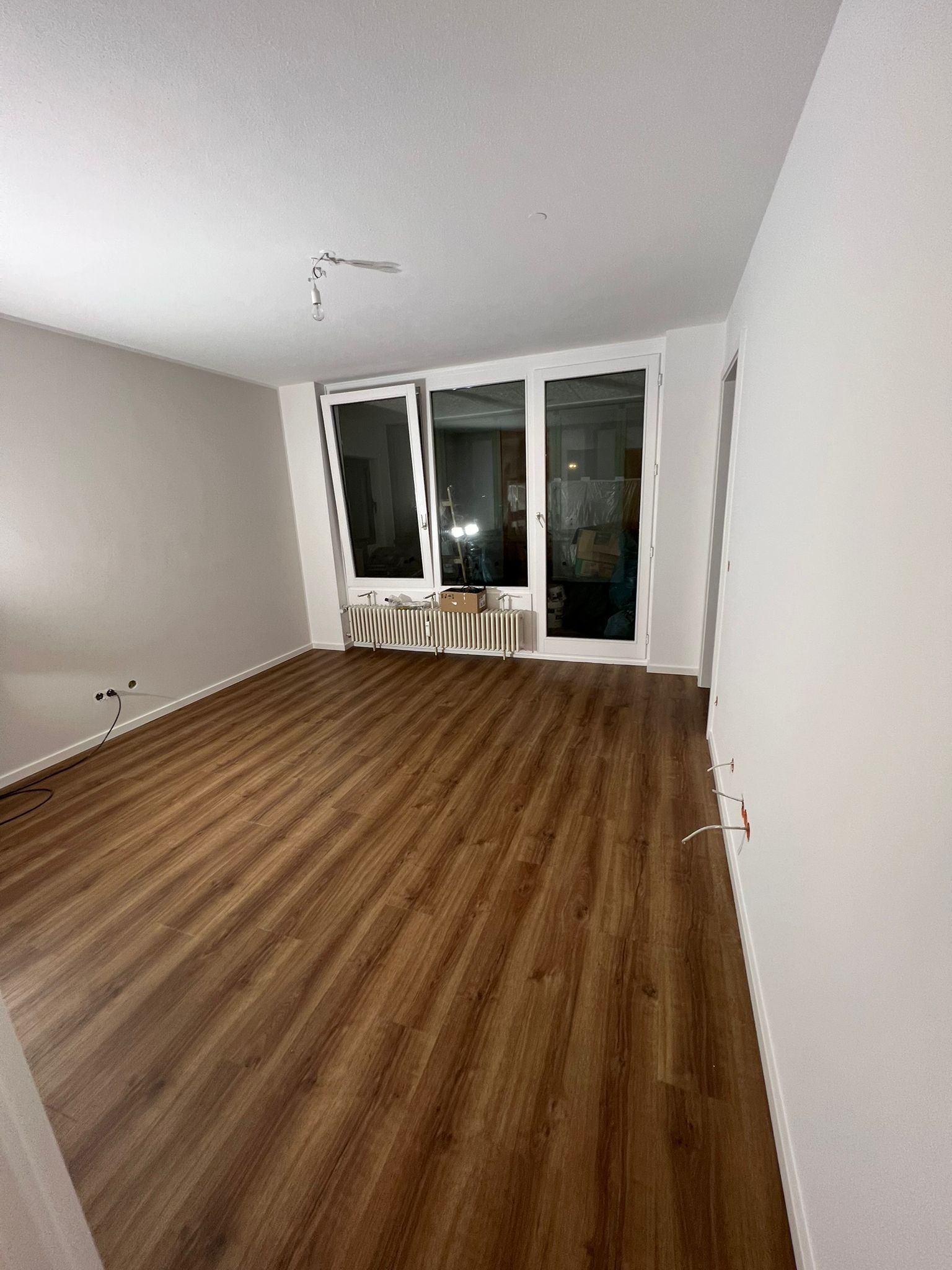 Pusty pokój z panelami podłogowymi w odcieniach brązu, świeżo pomalowane ściany na biało, widoczne gniazdka elektryczne i wystające kable, okno balkonowe z grzejnikiem pod spodem oraz wisząca...