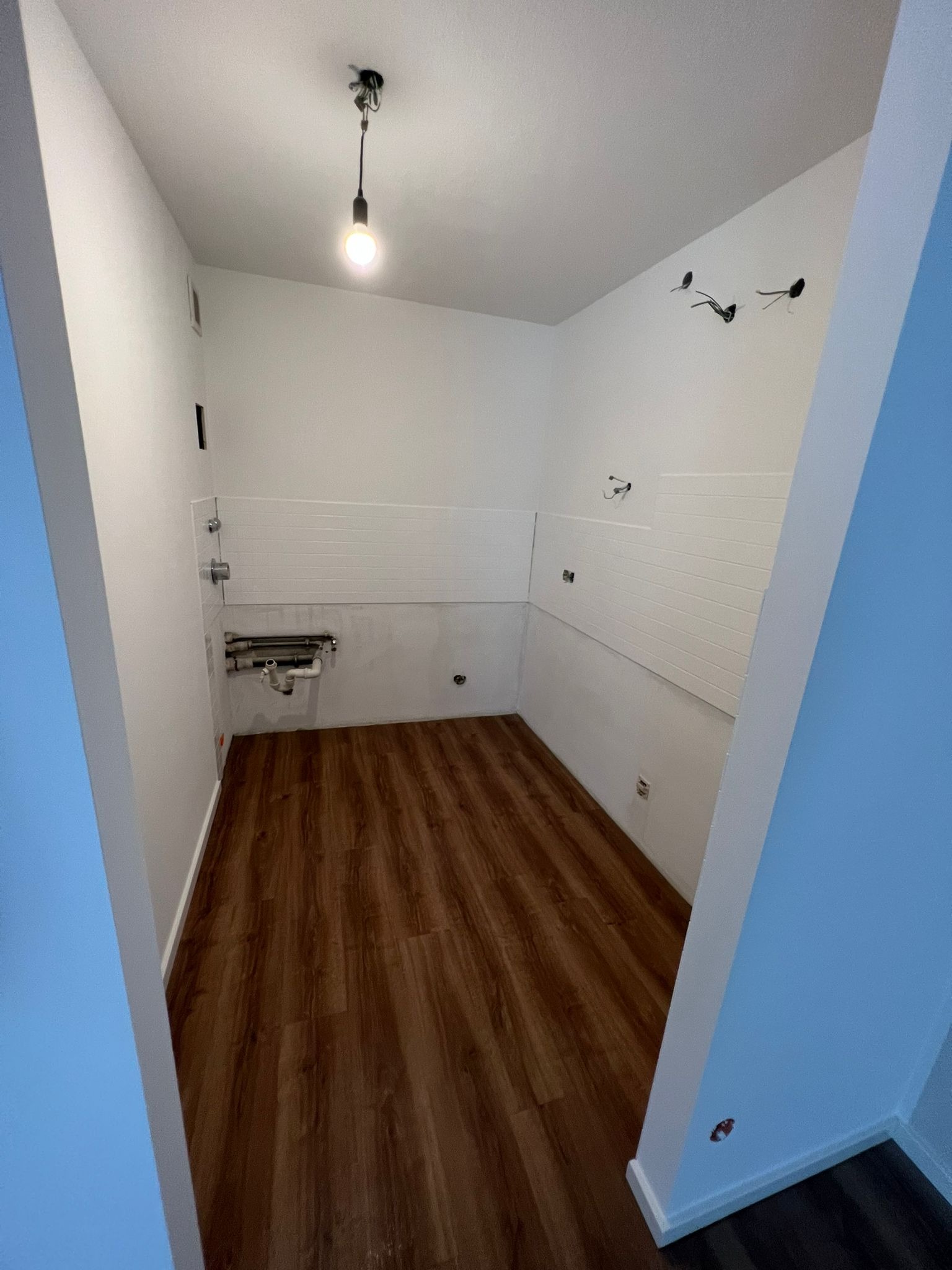 Wnętrze łazienki w trakcie remontu, widoczne nowe panele ścienne, drewnopodobna podłoga, instalacja wodno-kanalizacyjna oraz niedokończone punkty oświetleniowe.