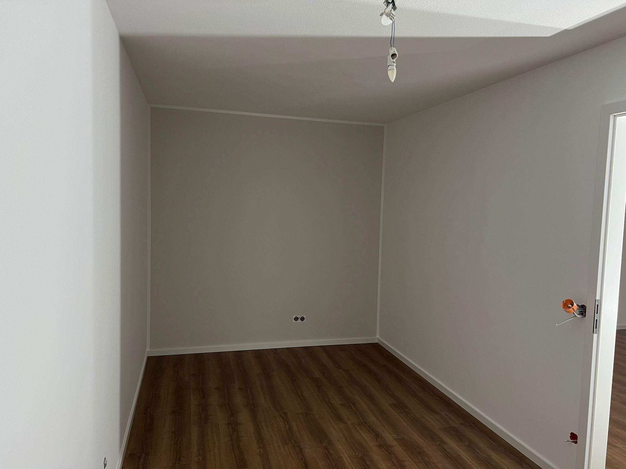 Pusty pokój po remoncie z panelami podłogowymi w odcieniach brązu, świeżo pomalowanymi ścianami na biało i szaro, widoczne gniazdka elektryczne, brakujące oprawy oświetleniowe i niedokończony...