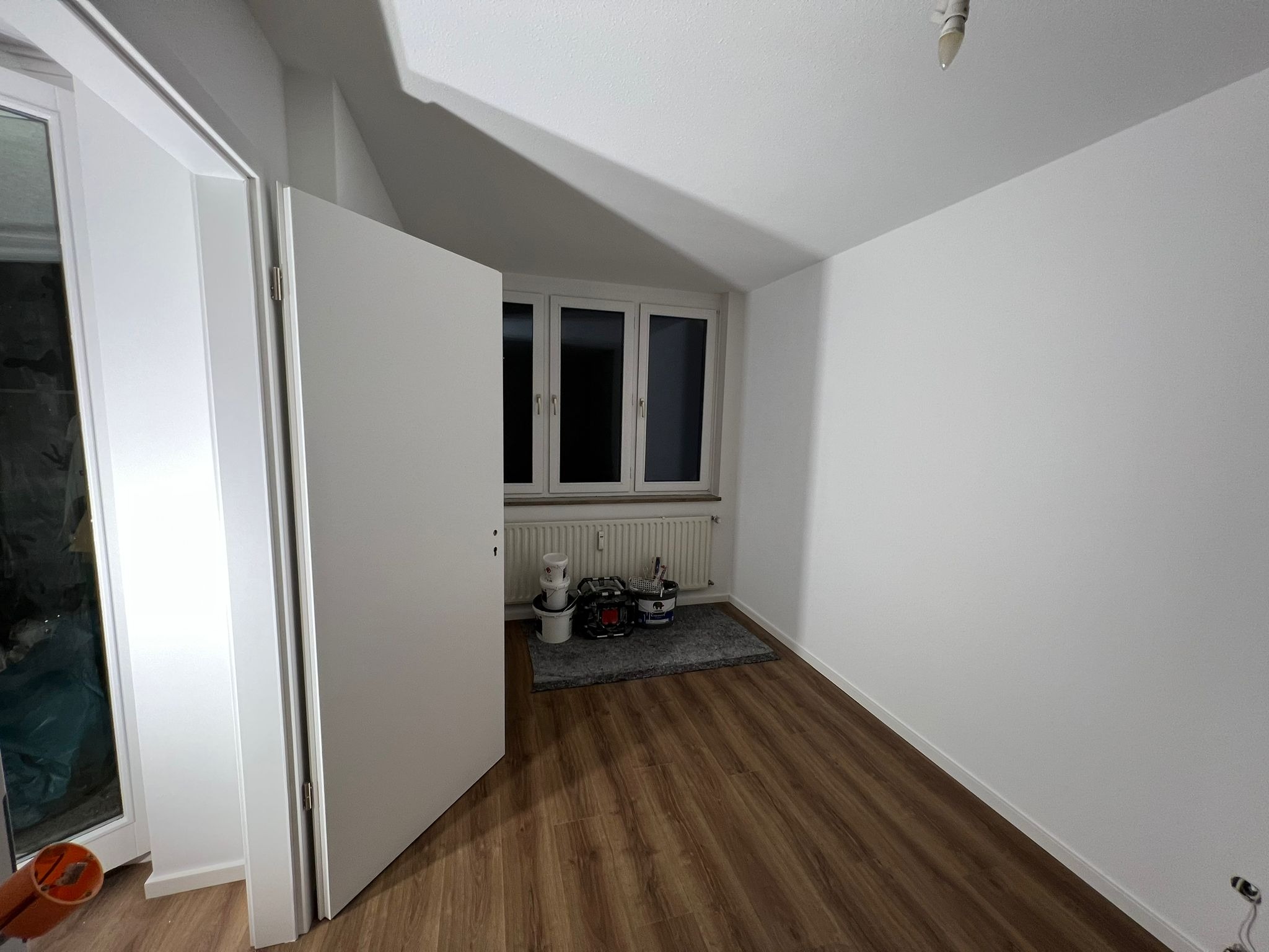 Pokój po remoncie z nową podłogą, świeżo pomalowanymi ścianami na biało i otwartymi białymi drzwiami. Widoczne narzędzia malarskie i puszki z farbą na dywaniku pod oknem.