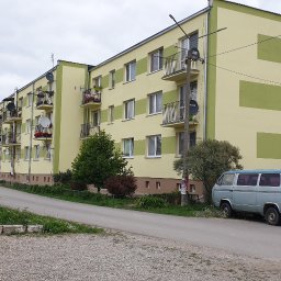 Norbert Gołombiewski BudNext - Żółto-zielony budynek mieszkalny z balkonami, numer 46, zaparkowany samochód dostawczy i dwa osobowe auta, widok z ulicy w pochmurny dzień.