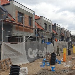 Norbert Gołombiewski BudNext - W trakcie budowy szeregu domów z czerwonymi dachami, widoczne rusztowania, ocieplenie ścian styropianem i prace wykończeniowe elewacji, teren budowy zabezpieczony siatką z logo firmy.