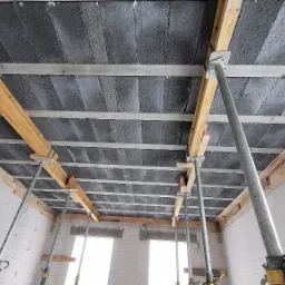 Wnętrze w trakcie budowy domu z widocznym stropem z betonowych bloczków, podpartym drewnianymi belkami i metalowymi stemplami, ściany z białych bloczków, dwa okna w tle.