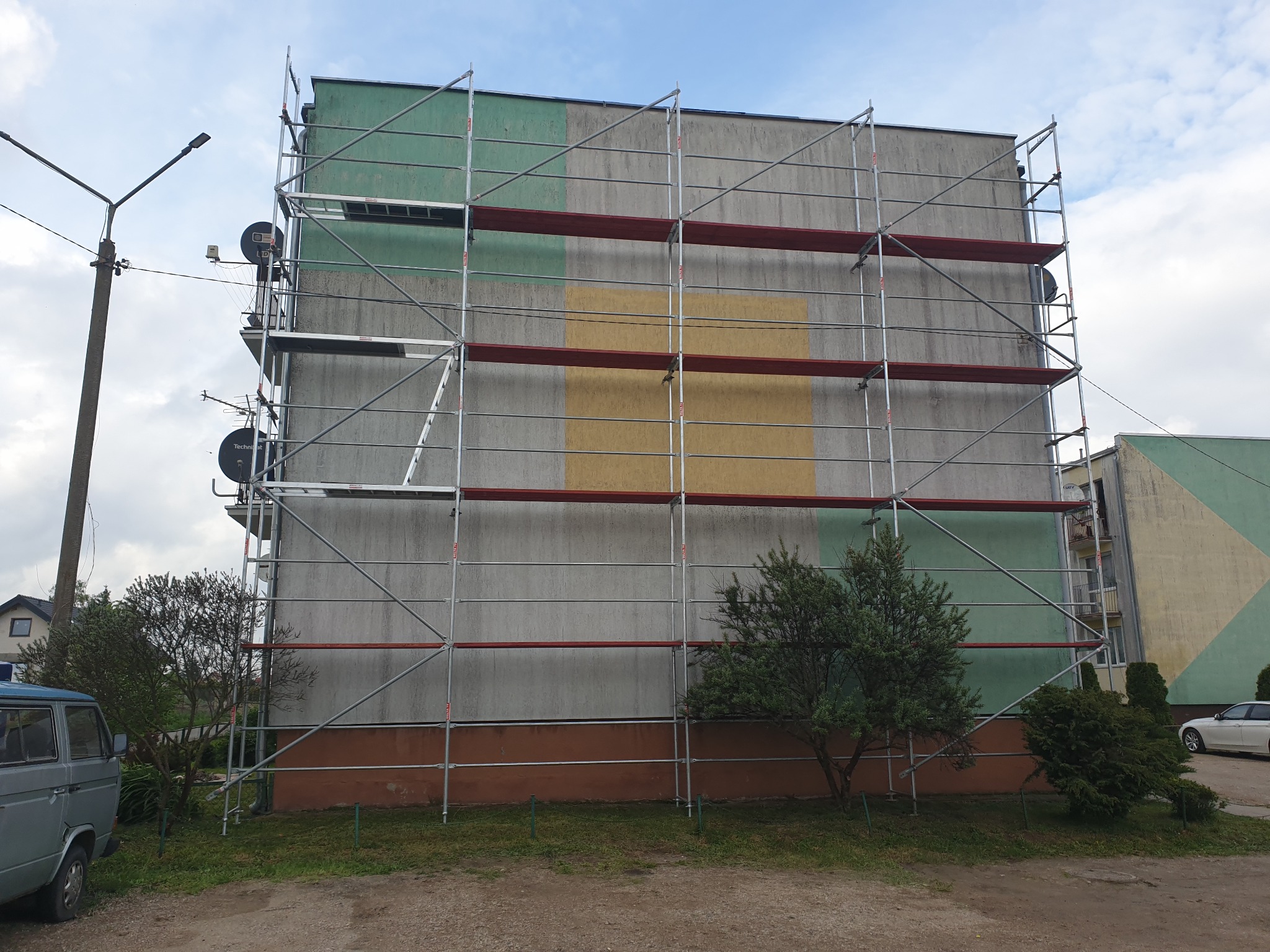 Budynek wielorodzinny w Olsztynie w trakcie prac elewacyjnych, pokryty rusztowaniem, z widocznymi fragmentami pomalowanej elewacji w odcieniach zieleni i żółci.