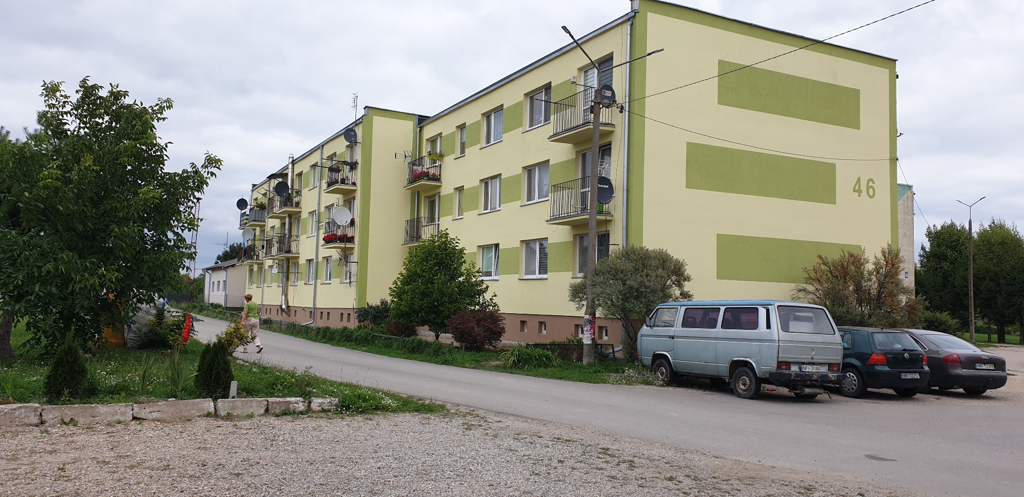 Żółto-zielony budynek mieszkalny z balkonami, numer 46, zaparkowany samochód dostawczy i dwa osobowe auta, widok z ulicy w pochmurny dzień.
