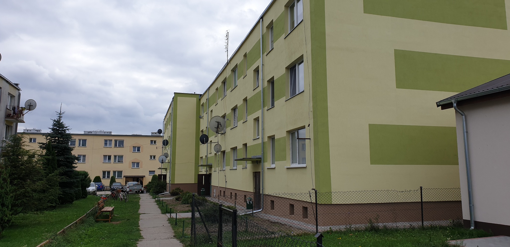 Blok mieszkalny po remoncie elewacji, z zielonymi pasami dekoracyjnymi, widoczne anteny satelitarne i chodnik.