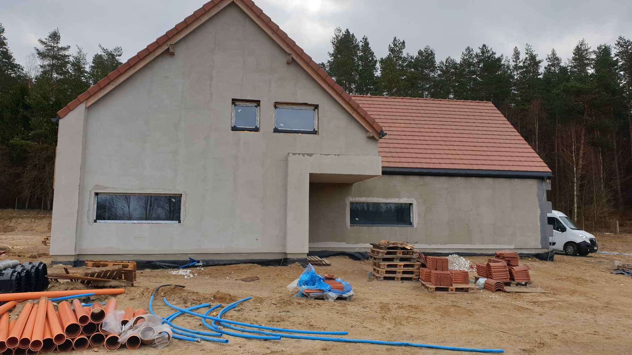 Surowy stan domu z szarą elewacją i czerwoną dachówką w trakcie budowy, widoczne okna bez parapetów, rury PCV i palety z dachówkami na piaszczystym gruncie, las w tle.