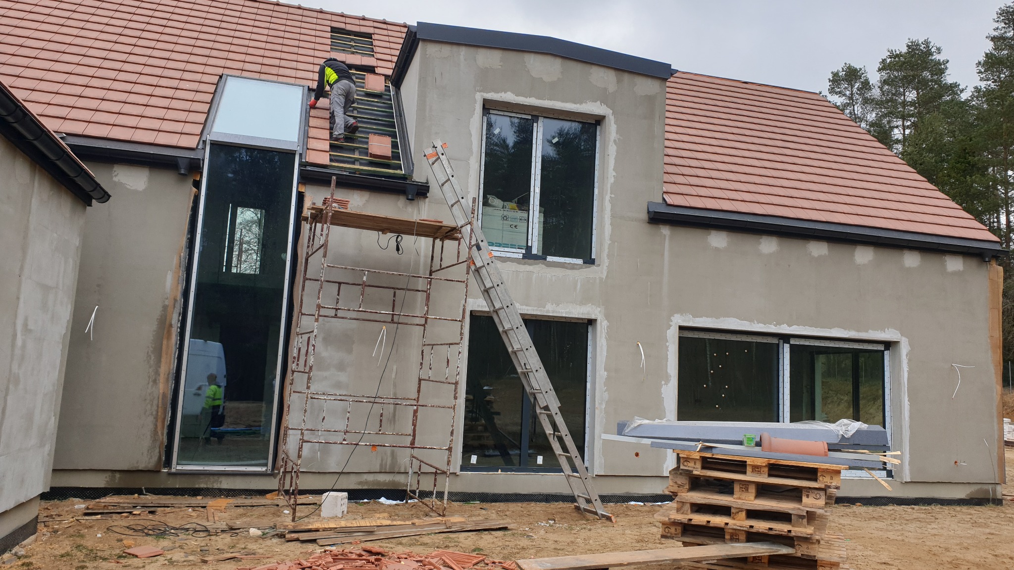 Budowa domu w stanie surowym: pracownik układa czerwoną dachówkę na dachu, widoczne rusztowania, drabina oparta o ścianę z oknami, palety z materiałami budowlanymi na ziemi.