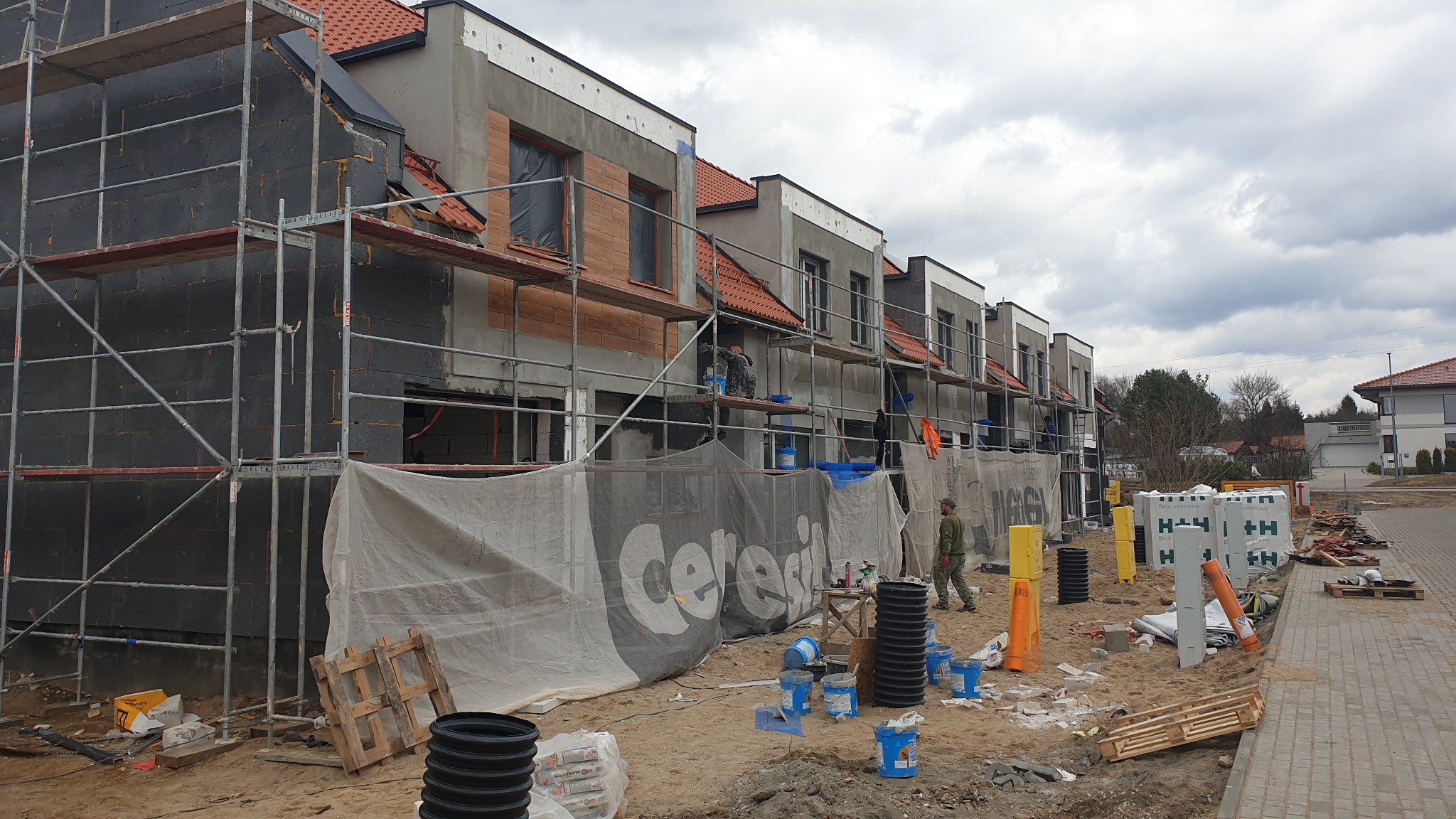 W trakcie budowy szeregu domów z czerwonymi dachami, widoczne rusztowania, ocieplenie ścian styropianem i prace wykończeniowe elewacji, teren budowy zabezpieczony siatką z logo firmy.