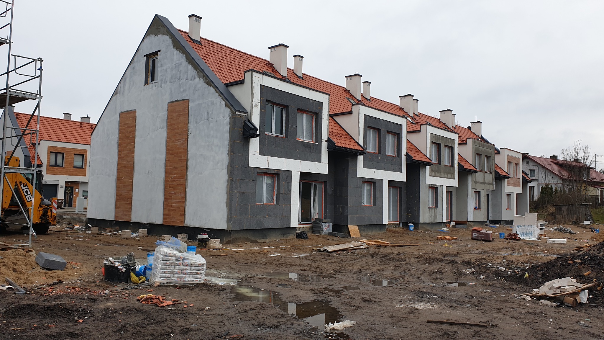 Szeregówka w trakcie budowy: część budynków z szarą elewacją i czerwonym dachem, inne z ociepleniem grafitowym, widoczny sprzęt budowlany i materiały na błotnistym terenie.
