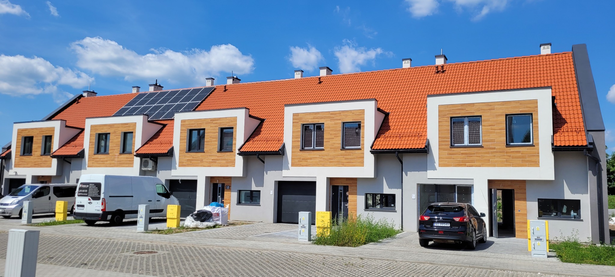 Nowoczesne segmenty mieszkalne z panelami słonecznymi na dachu, wykończone elewacją w odcieniach bieli, brązu i szarości, z zaparkowanymi samochodami i furgonetką firmy budowlanej na podjeździe.