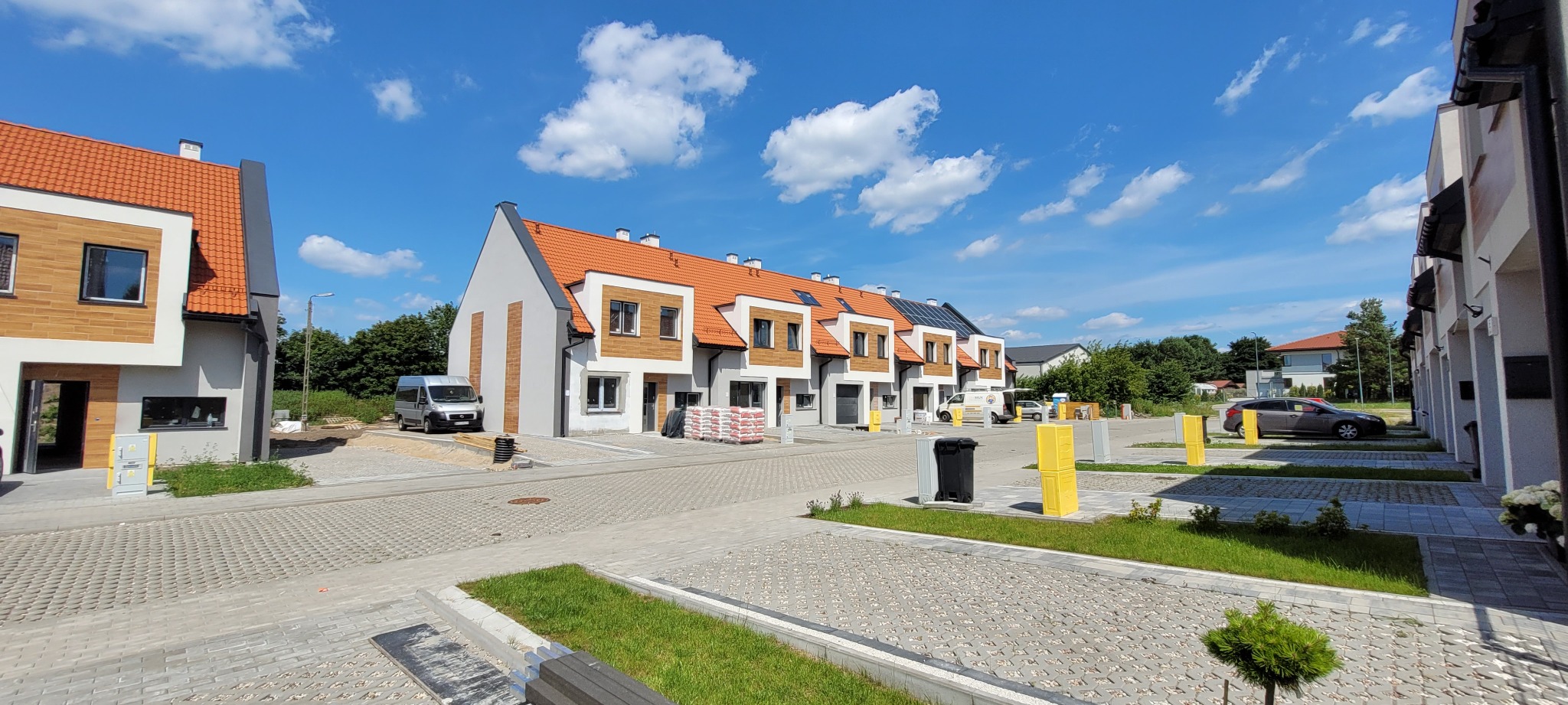 Nowo wybudowane szeregówki z pomarańczową dachówką, jasną elewacją i drewnianymi akcentami, przed jednym z domów leżą worki z materiałami budowlanymi, na podjeździe stoi bus.