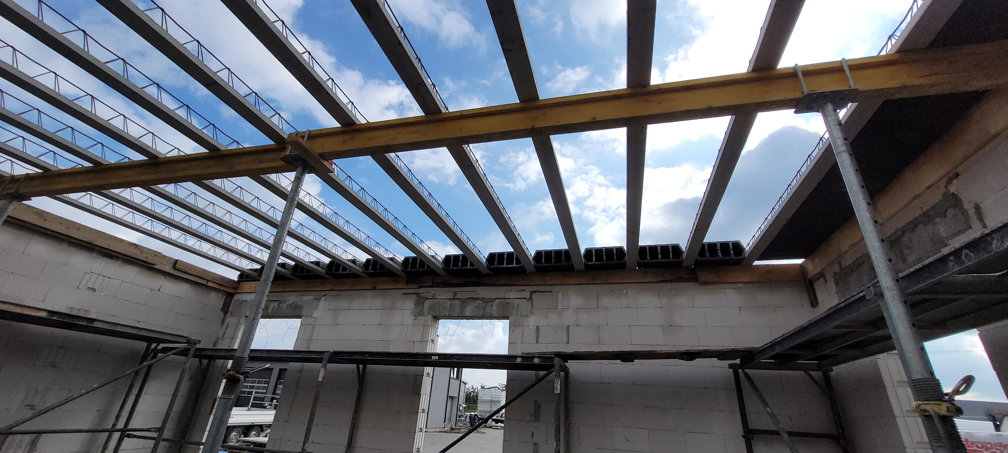 Konstrukcja stropu żelbetowego w trakcie budowy domu z bloczków, widoczne szalunki, stalowe podpory i fragment nieba przez otwory w konstrukcji.