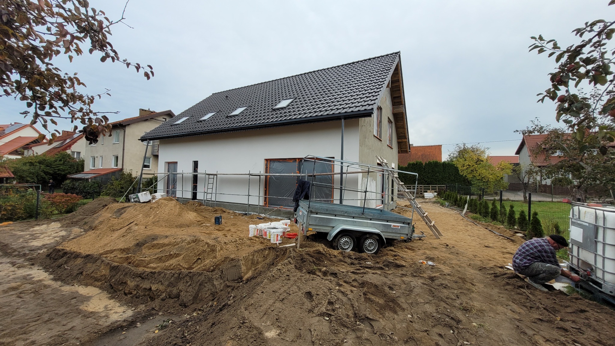 Nowo wybudowany dom z ciemną dachówką, częściowo otynkowany, z rusztowaniem, przyczepką i stertą piasku na posesji w trakcie prac wykończeniowych.